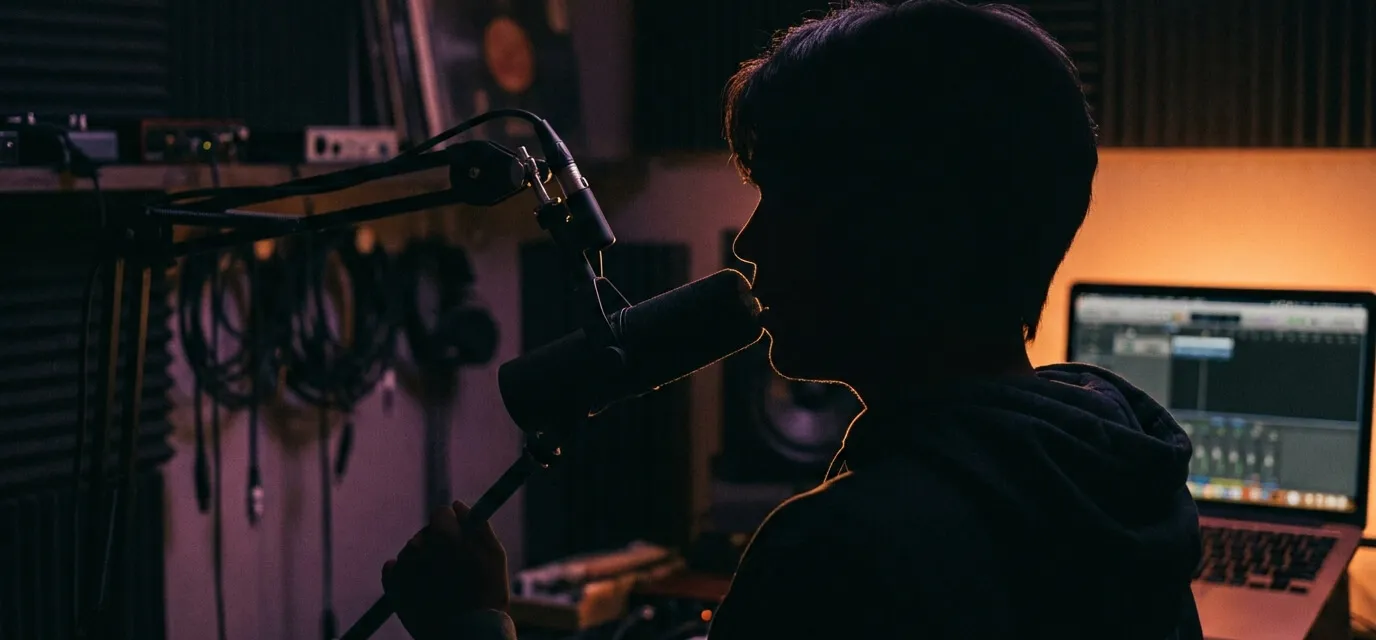 Artist singing into a studio microphone with a DAW visible on a laptop in the background